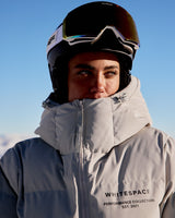 A close-up of a snowboarder wearing the WHITESPACE Cropped Puffer Jacket (Pigeon) and WHITESPACE Magnetic MD Icon Goggles (White) against a clear blue sky