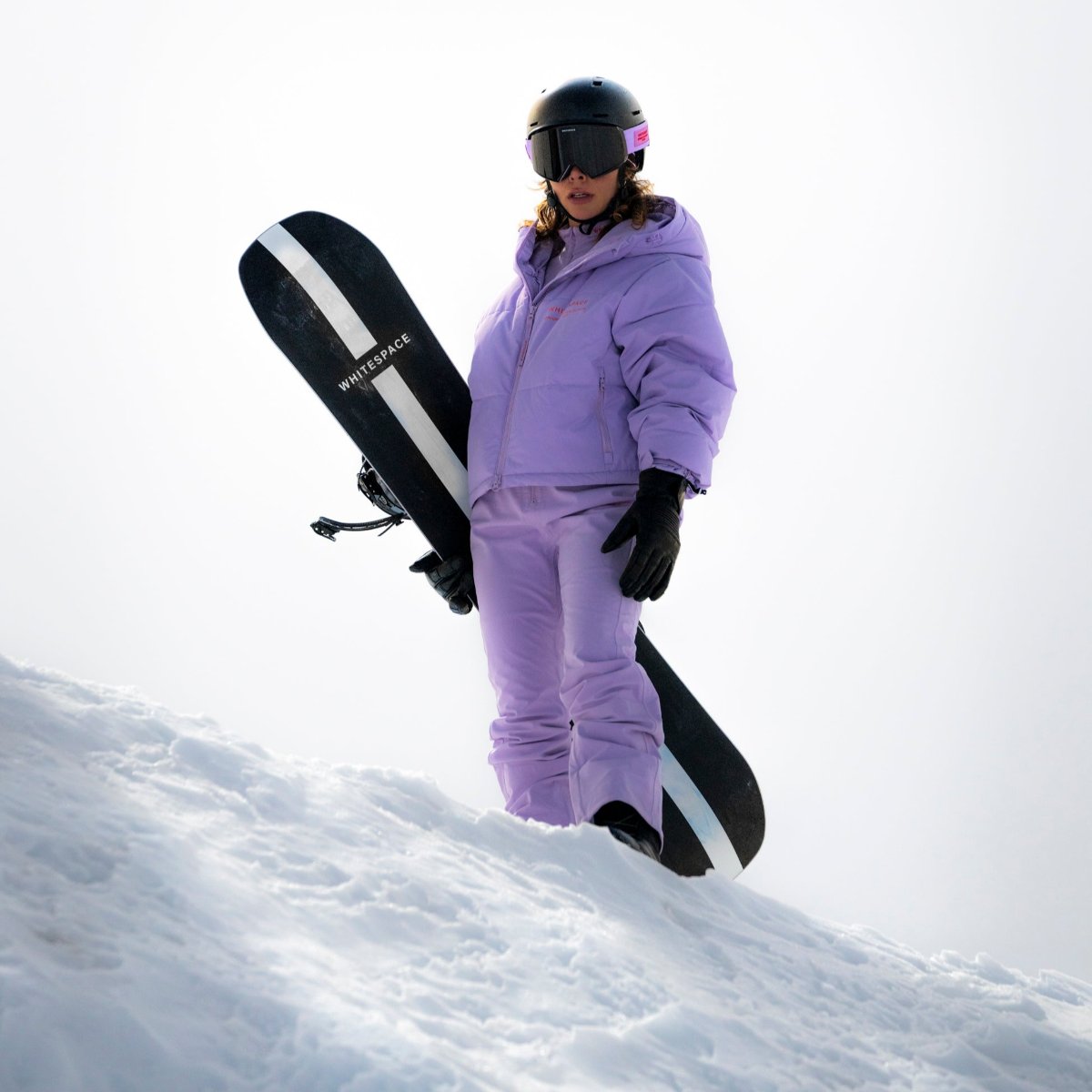 Woman standing on a snowy mountain holding a WhiteSpace snowboard and wearing Cropped Puffer Jack in Lilac Purple and pants.