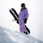 Woman standing on a snowy mountain holding a WhiteSpace snowboard and wearing Cropped Puffer Jack in Lilac Purple and pants.