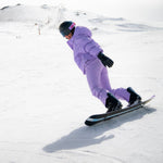 Woman snowboarding down a snowy mountain wearing Cropped Puffer Jack in Lilac Purple and pants.