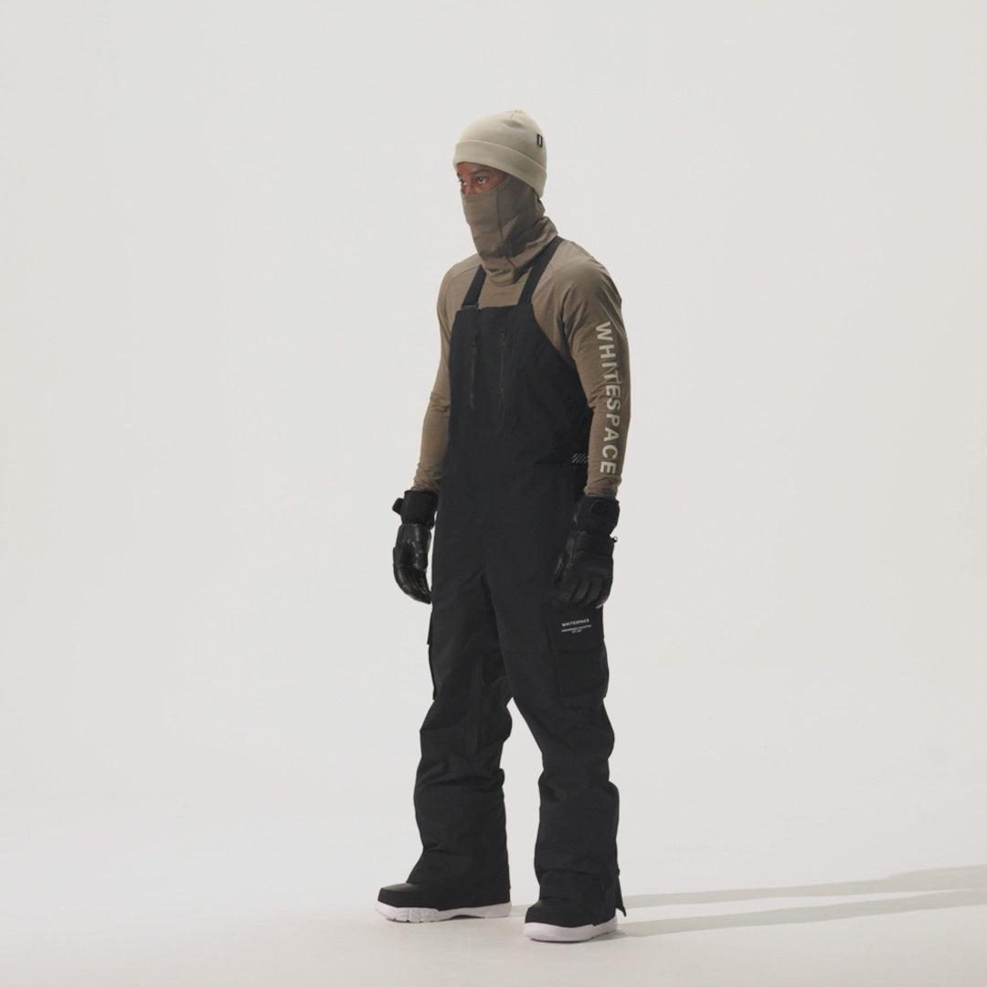 Video of model wearing 2L Insulated Cargo Bib Pants in Black over a beige shirt and gaiter, on a white background.