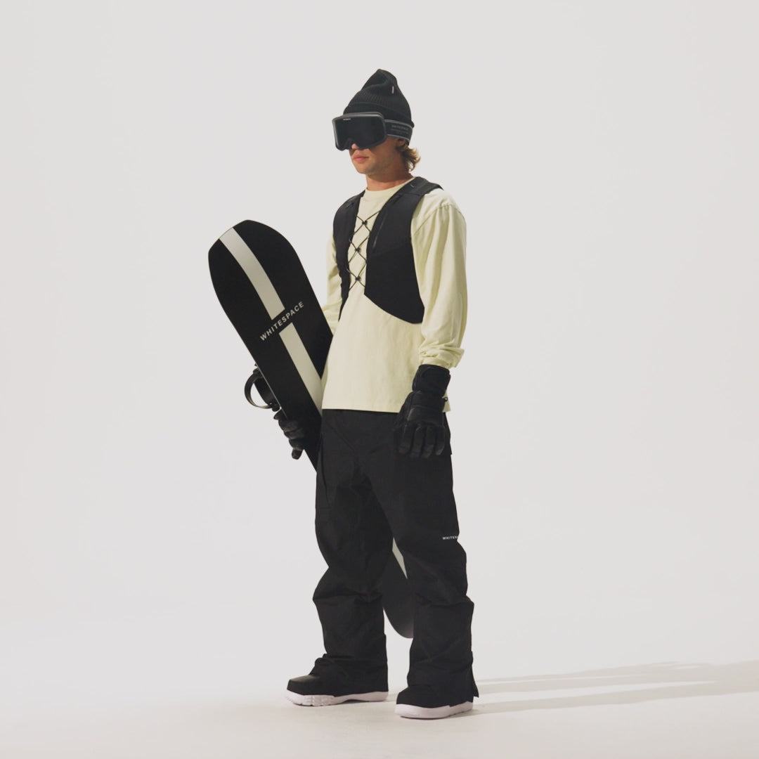 Video of model holding a snowboard and wearing a  REC Cargo Vest in Black, black pants, and a cream shirt on a white background.