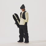 Video of model holding a snowboard and wearing a  REC Cargo Vest in Black, black pants, and a cream shirt on a white background.