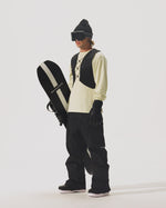 Video of model holding a snowboard and wearing a  REC Cargo Vest in Black, black pants, and a cream shirt on a white background.