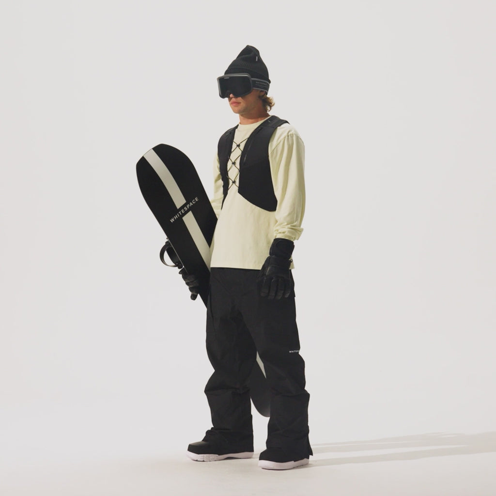 Video of model holding a snowboard and wearing a  REC Cargo Vest in Black, black pants, and a cream shirt on a white background.